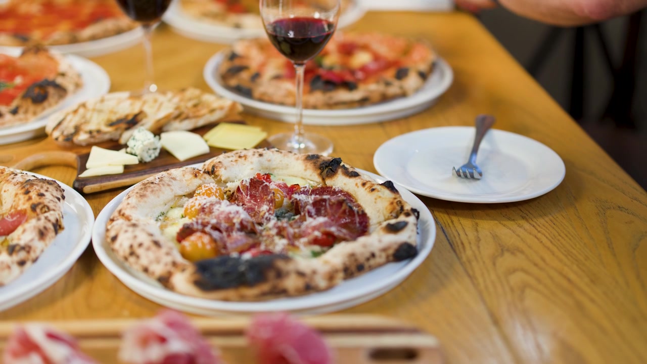 Primo piano di mani che tagliano pizza artigianale con vino rosso e salumi su tavolo di legno.