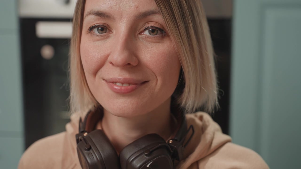 White woman music lover with headphones, kitchen closeup of glass on floor, listener reaches for water between tracks, musician nods to beat while sipping, portrait smile and candid eye contact, cozy