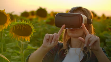 the girl farmer manager with long hair in plaid shirt and jeans is working in vr glasses. she is engaged in the working process. it is enjoyable sunny evening in the sunflower field at sunset.