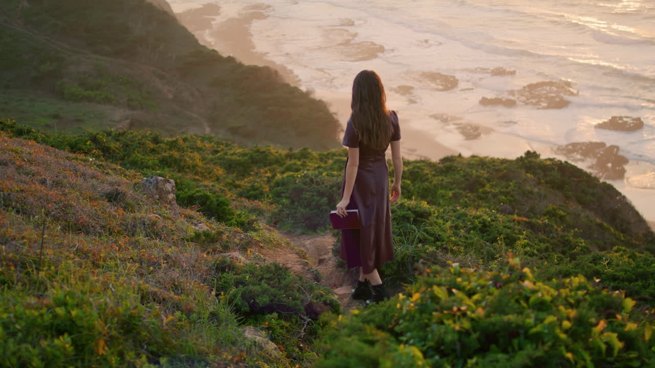 Donna sconosciuta che cammina su una collina vicino all'oceano tenendo un libro. La donna si gode la costa di sera.