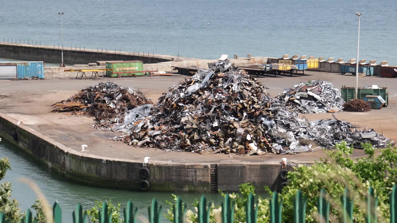 Pilha de sucata metálica no cais do Porto de Seaham, vista aproximada de aço e alumínio torcidos misturados em uma área de reciclagem de porto industrial com muro de contenção e contêineres ao fundo.