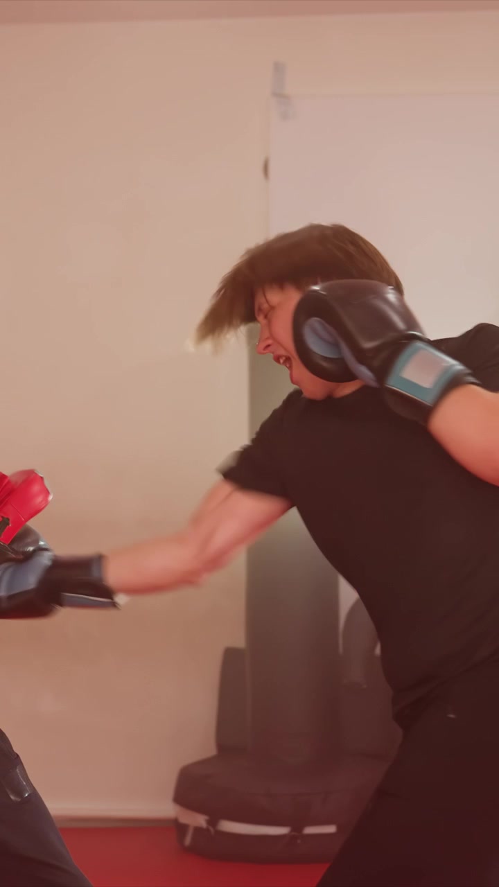 Giovani uomini praticano tecniche di boxe in una palestra al coperto. Un uomo sferra pugni verso dei colpitori rossi tenuti. L'ambiente presenta uno sfondo a parete semplice con attrezzi da esercizio visibili.