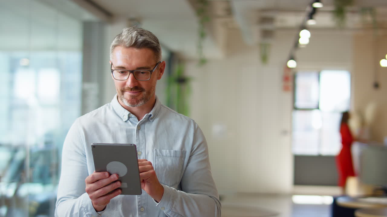 Mature Businessman Standing In Modern Open Plan Office Using Digital Tablet