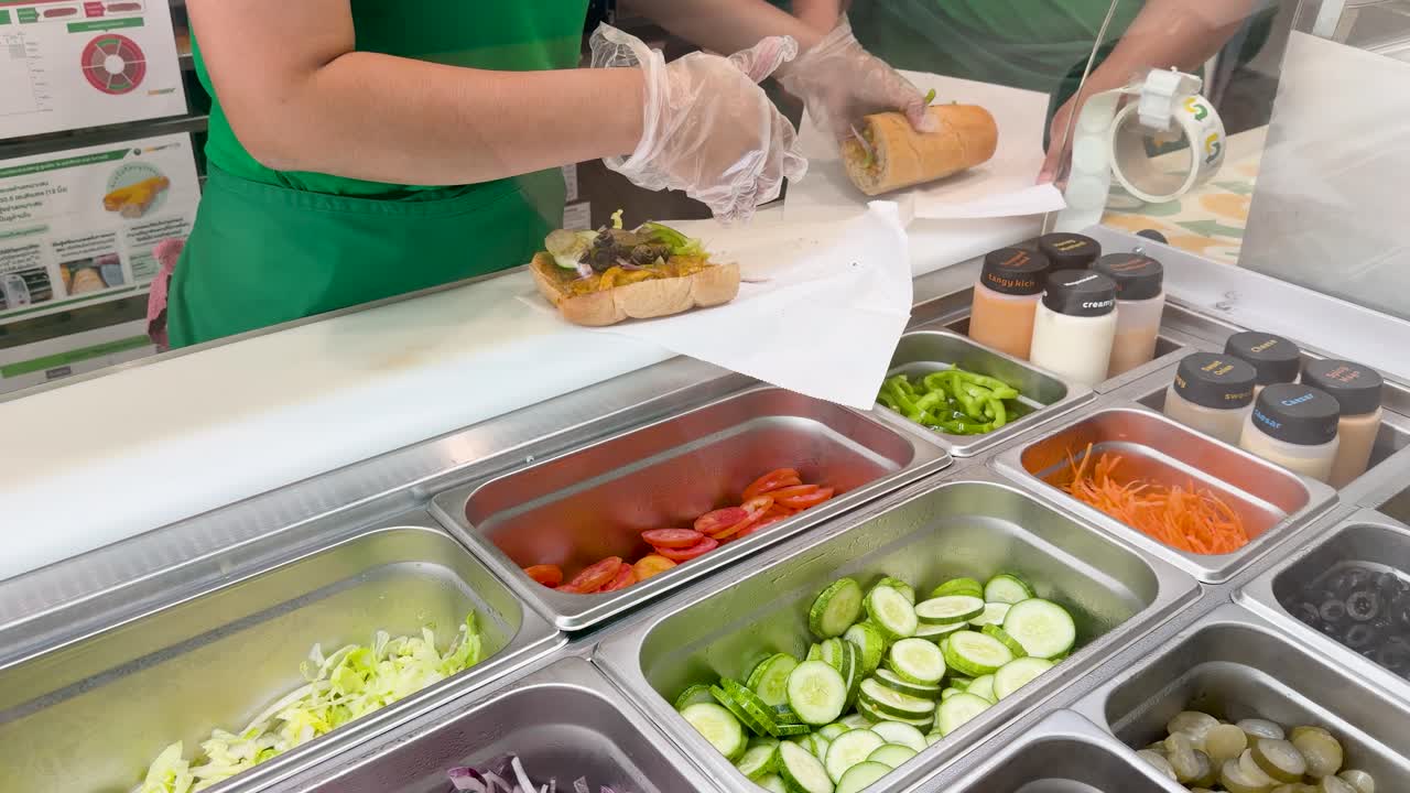 Un addetto alla ristorazione con i guanti assembla accuratamente un fresco panino sub con verdure.