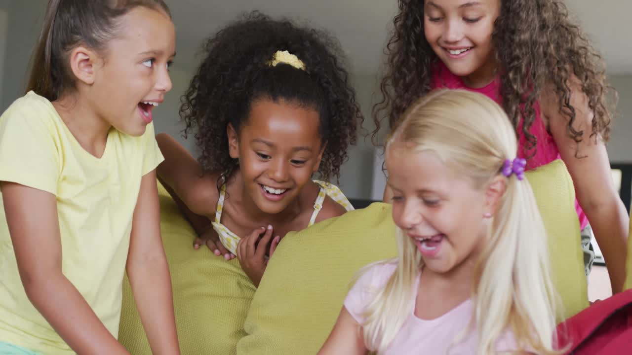 Video of diverse girls sitting at school common room with tablet, talking and laughing