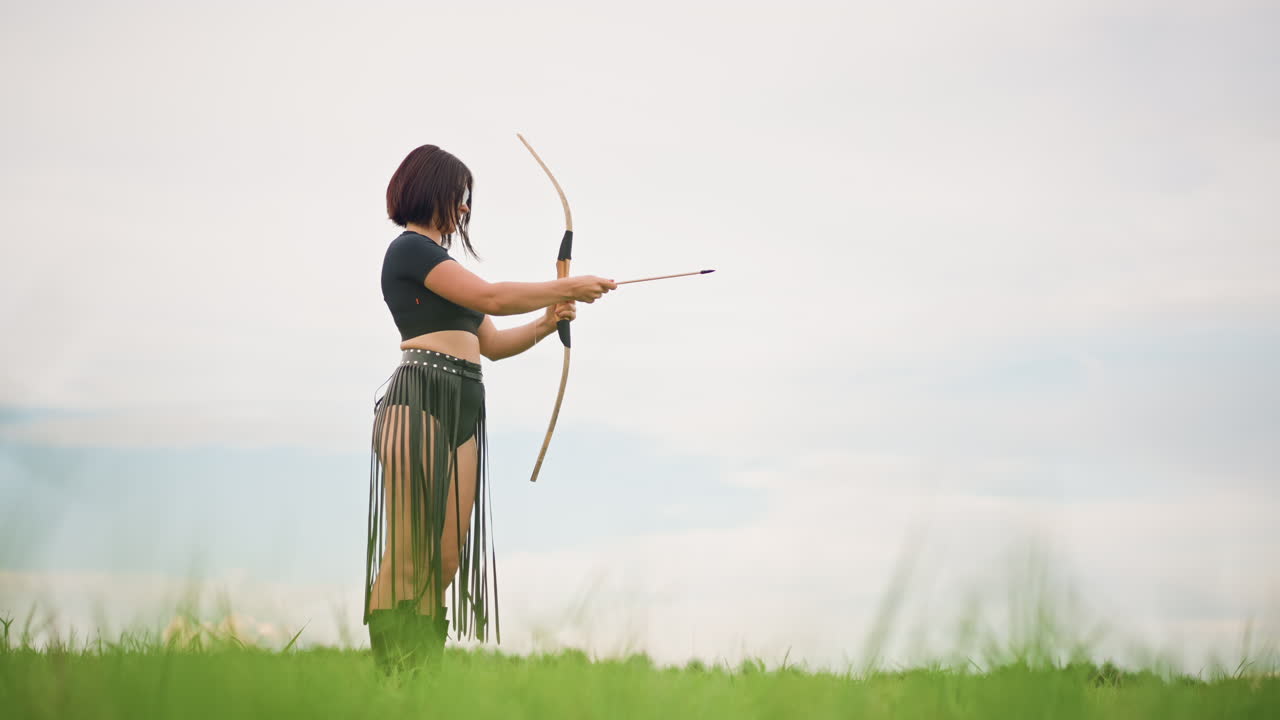 Una donna arciere in piedi in una prateria, che indossa una gonna di pelle, tende un arco di legno puntando con mira ferma verso l'orizzonte lontano, cielo nuvoloso e delicato, erba alta e verde in primo piano, atmosfera cinematografica e solitaria che trasmette