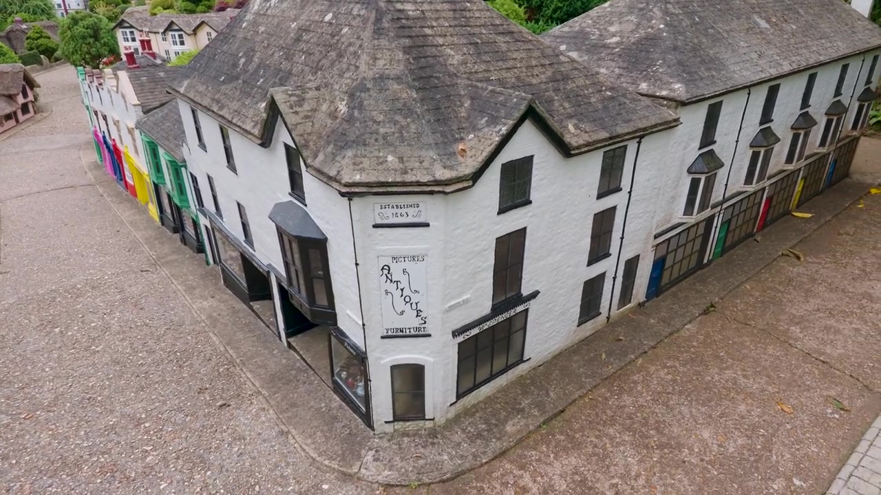 Modellini di negozi colorati al Godshill Model Village mentre la telecamera si sposta a sinistra, poi torna indietro lungo una strada in miniatura di edifici dettagliati e vetrine, evidenziando un'affascinante esposizione turistica