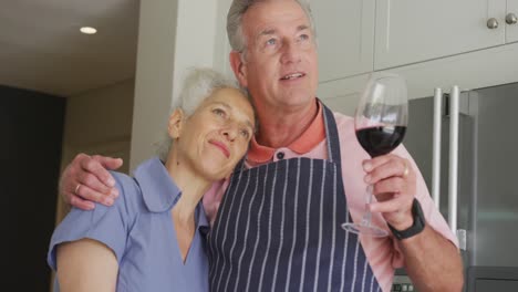 Happy-caucasian-senior-couple-wearing-aprons-embracing-and-drinking-wine