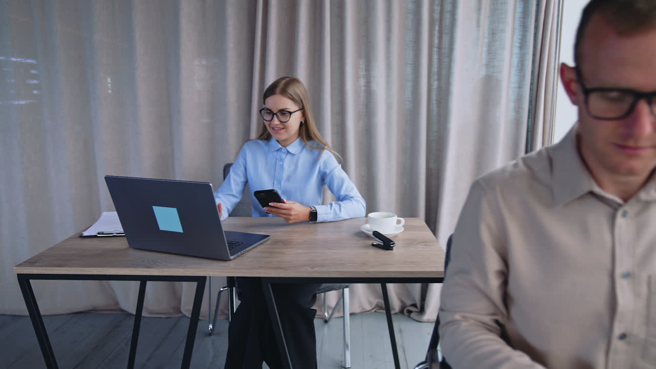 Blonde lady office employee uses phone and laptop at once. Male colleague opens his paper notebook to takes notes. Office background.