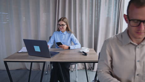 blonde lady office employee uses phone and laptop at once. male colleague opens his paper notebook to takes notes. office background.