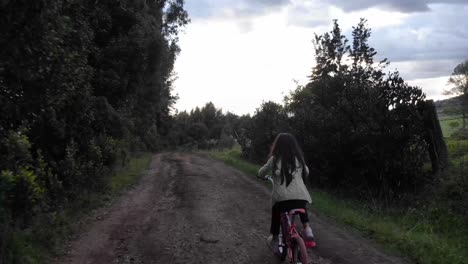 Toma-De-Seguimiento-De-Una-Niña-Montando-Su-Bicicleta-Por-Un-Camino-De-Tierra-Irregular