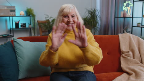 Abuela-Mujer-Sonriendo-Amistosamente-A-La-Cámara-Saludando-Con-La-Mano-Haciendo-Un-Gesto-De-Saludo-En-Casa-En-El-Sofá