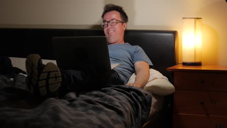 man laying upright in bed gets excited watching sport on his laptop