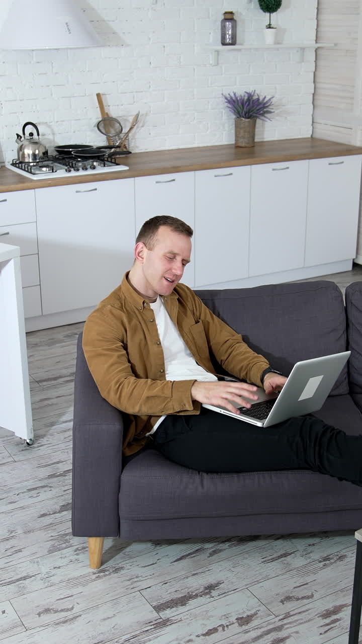 Relaxed guy working on a wireless computer at home. Young man laying on a sofa and typing on a laptop on modern kitchen background. Job at quarantine. Vertical video