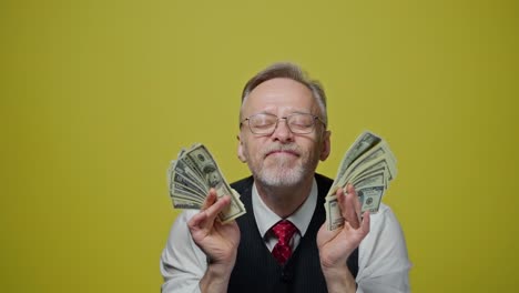 portrait of grey-haired man with dollars. smiling mature businessman in glasses waving dollar bills and touches his face with money happily. positive emotions.