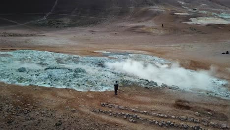 aerial, orbit, drone shot, around a woman taking pictures of a steaming crater emitting sulfuric gas, at hverir geothermal area, on a sunny, summer day, in north iceland