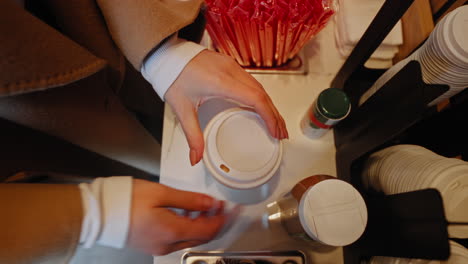 close up of a young woman hands closing the plastic lid on a coffee paper cup, inside a coffee shop, slow motion