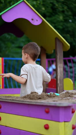 young boy playing in a sandbox with sand touching it with his hands. kid turns around and throws some sand up. vertical video.