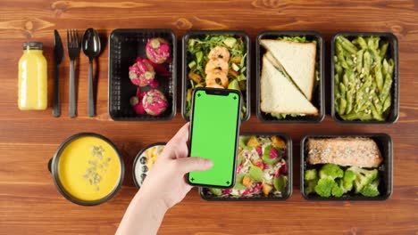 food delivery top view, take away meals in disposable containers on wooden table. lunch boxes with cooked vegetarian dishes, using phone with chroma green screen. healthy diet. catering service