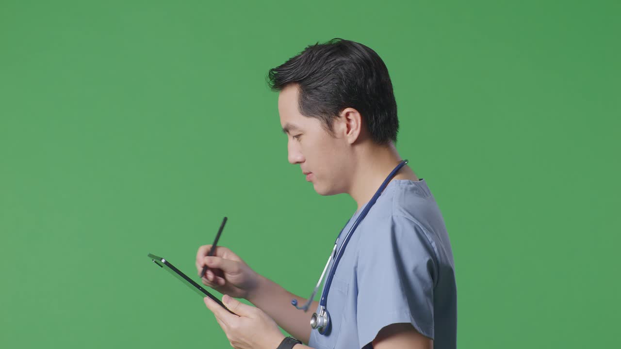 측면 뷰의 가까운 아시아 남성 의사는 스테토스코프를 가지고 병원에서 녹색 화면 배경에서 는 동안 태블릿에 메모를 취합니다.