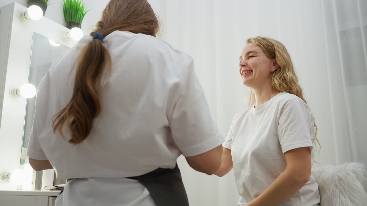 Client with long blond hair smiles while looking into mirror and engaging with beautician wearing gloves and mask in bright salon with vanity lights, white curtain, and cozy fur chair