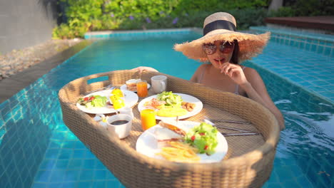 Asian-woman-takes-a-bite-from-her-breakfast-on-a-floating-tray-while-in-a-swimming-pool