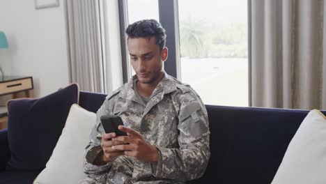 thoughtful biracial male soldier in uniform sitting on sofa using smartphone, slow motion