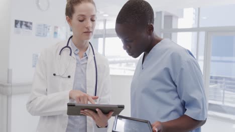 diverse female doctor and nurse looking at tablets and talking in hospital corridor, in slow motion