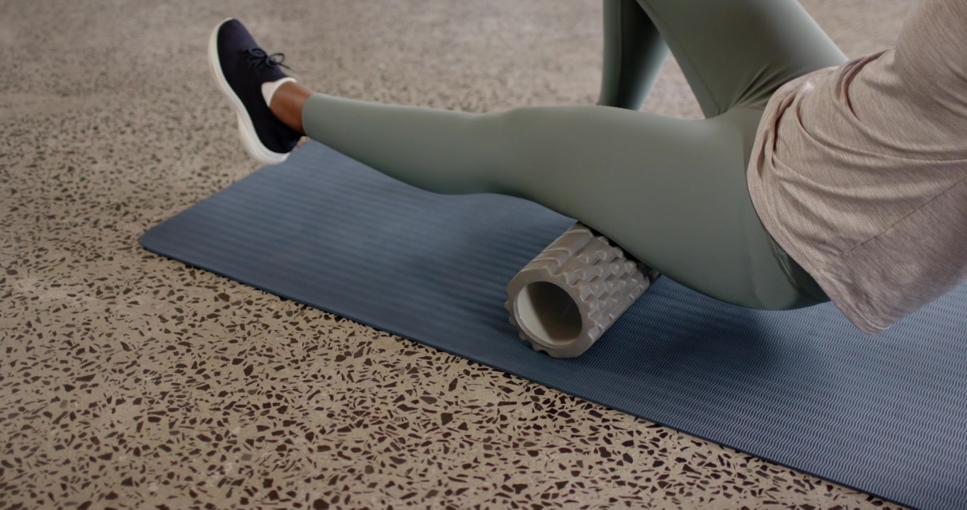 African American woman placing foam roller under right thigh rolling for relief on blue mat