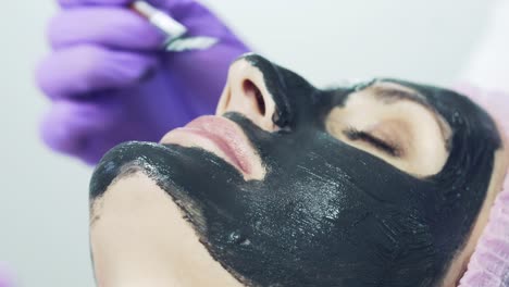 pretty woman is getting a facial treatment with a black mask at the health spa. beautician applying a mask of the therapeutic black mud. close-up