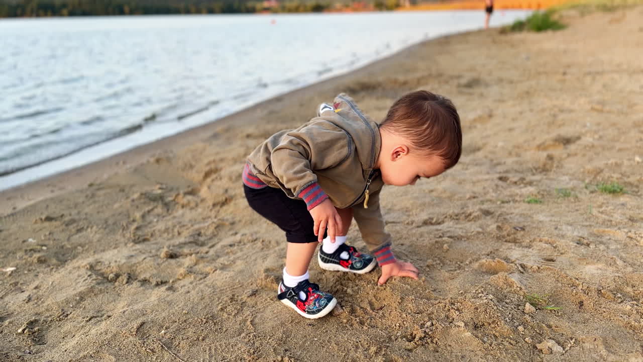 Simpatico bambino caucasico sulla riva del fiume. Il bambino getta la sabbia dalla riva nell'acqua.
