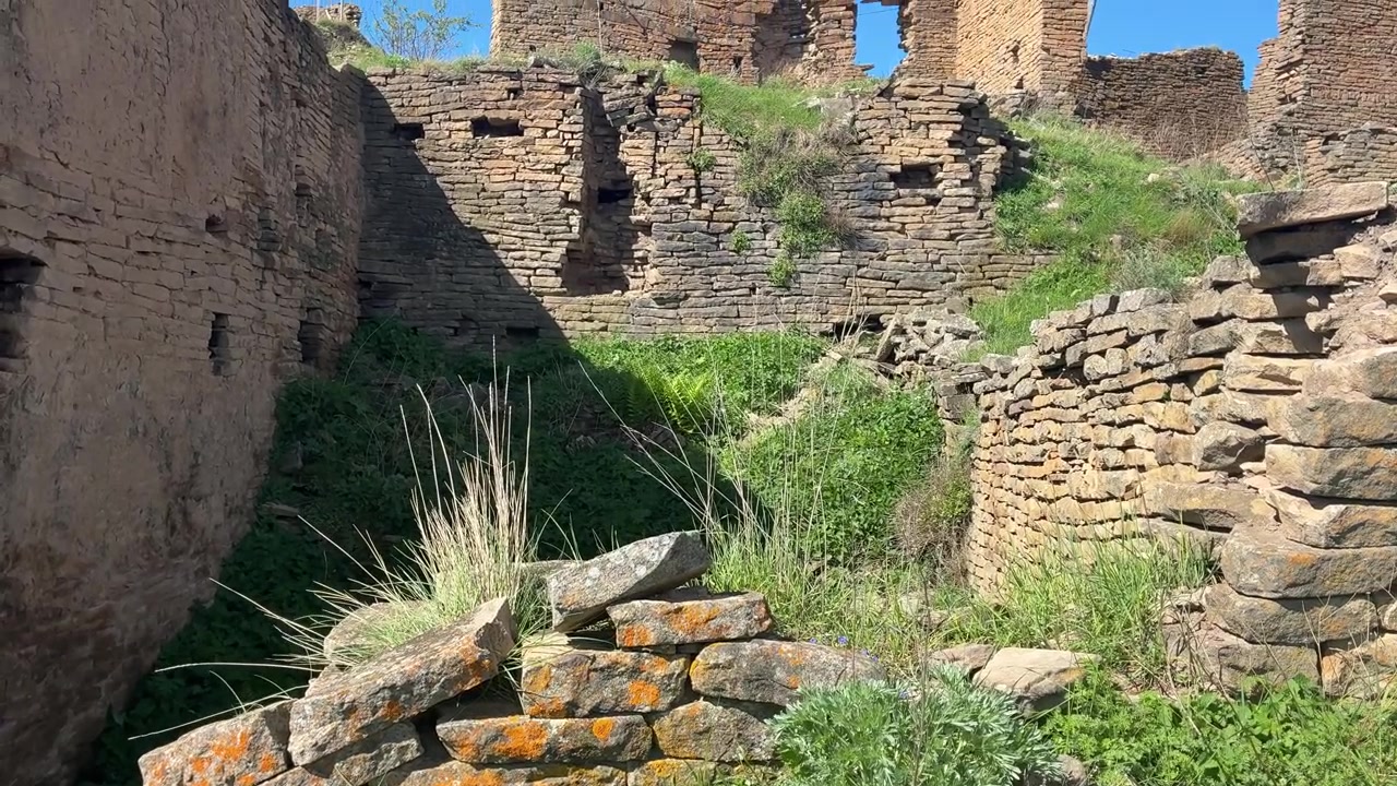 Ujęcie z niskiej perspektywy powoli panoramuje w górę, ukazując starożytne kamienne ruiny wioski Kakh, z zieloną trawą rosnącą wśród rozpadających się murów pod czystym, błękitnym niebem.