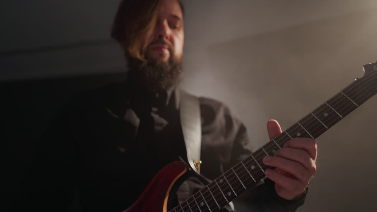 Bearded guitarist in a black shirt passionately playing a red guitar, subtly moving his head in a softly lit room