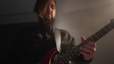 guitarrista barbudo con una camisa negra tocando apasionadamente una guitarra roja, moviendo sutilmente la cabeza en una habitación suavemente iluminada