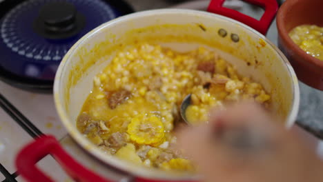 a chef serves a thick and corn-based locro argentino dish from a red pot on the stove.
