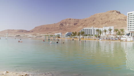 turistas adultos con psoriasis están flotando y disfrutando del sol caliente del mar muerto con hoteles en el fondo