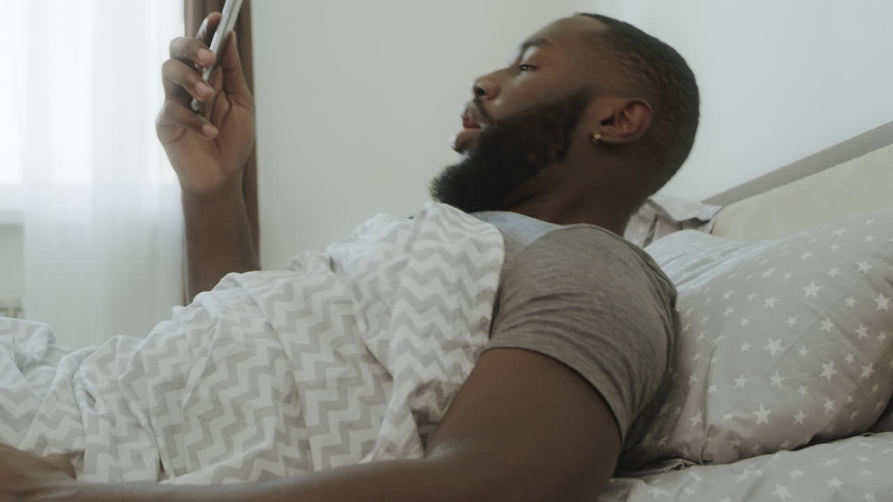 Uomo nero scioccato seduto a letto. Lo smartphone gli cade di mano.