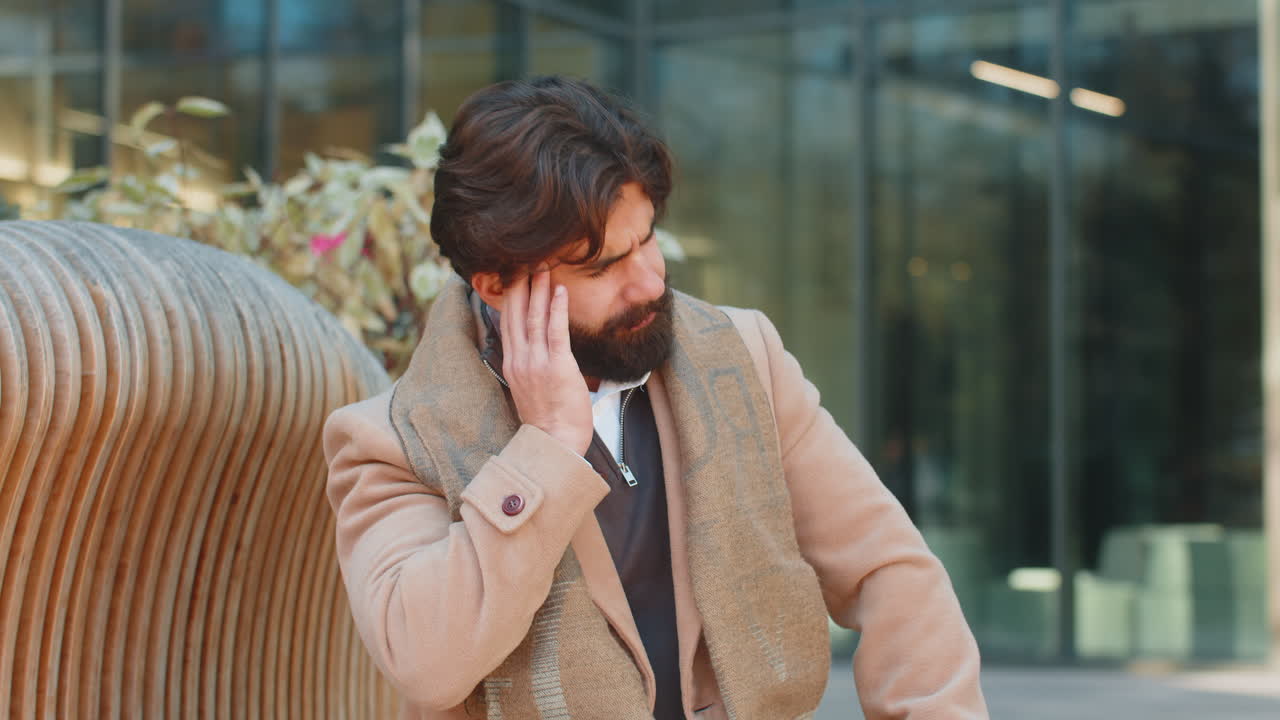 Sick young indian business man feels headache while sitting against building on city street