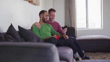 Multi-ethnic-gay-male-couple-sitting-on-couch-looking-at-smartphone-together