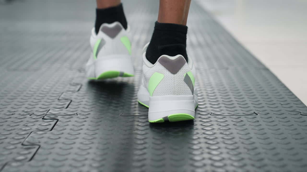 close up rear view of white neon green running shoes walking on textured black gym floor mat under bright overhead lights in spacious corridor with blurred equipment and windows beyond