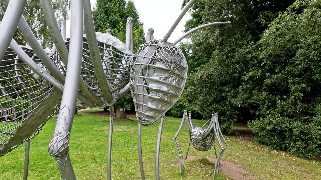A smooth camera move advances toward a large metallic insect sculpture set on green grass in a lush botanical garden under diffuse daylight
