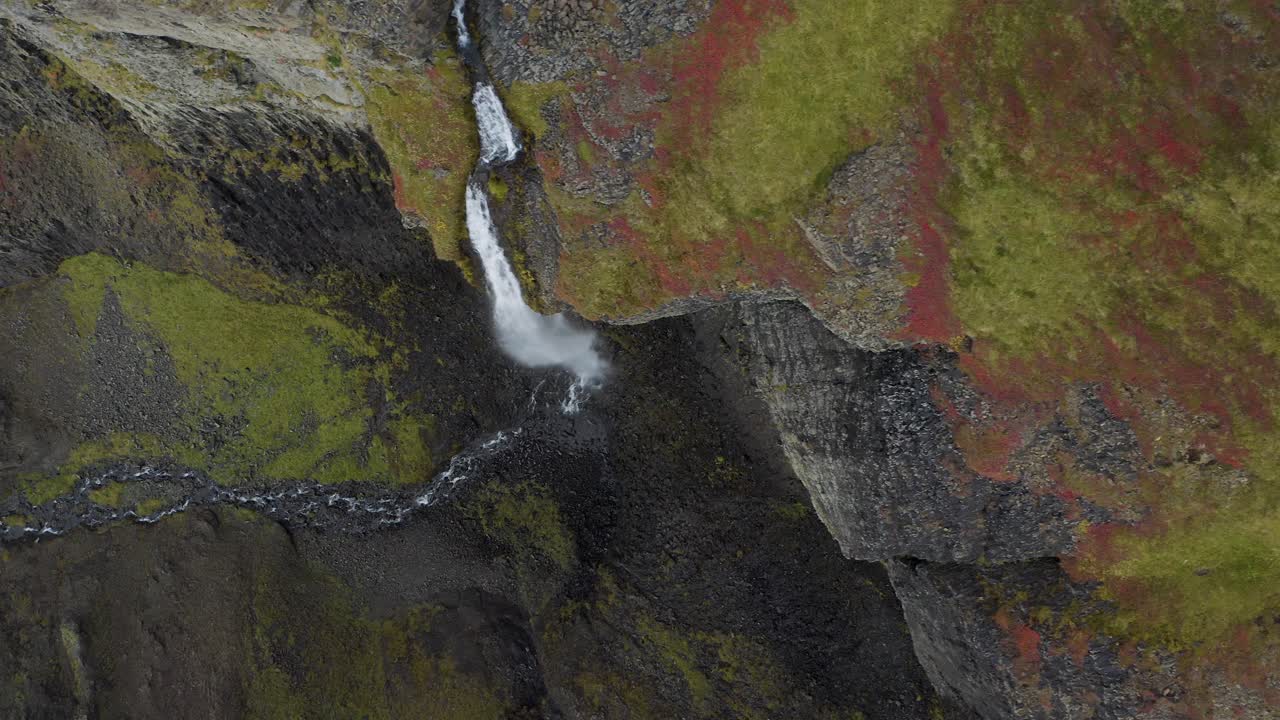 toma aérea de arriba hacia abajo de una cascada que cae a lo largo de montañas rocosas de musgo en la isla de islandia - toma panorámica