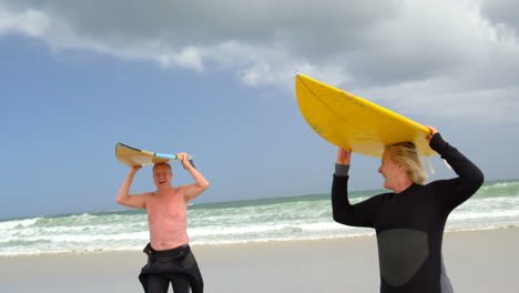 vista frontal de una vieja pareja caucásica llevando una tabla de surf en la cabeza en la playa 4k