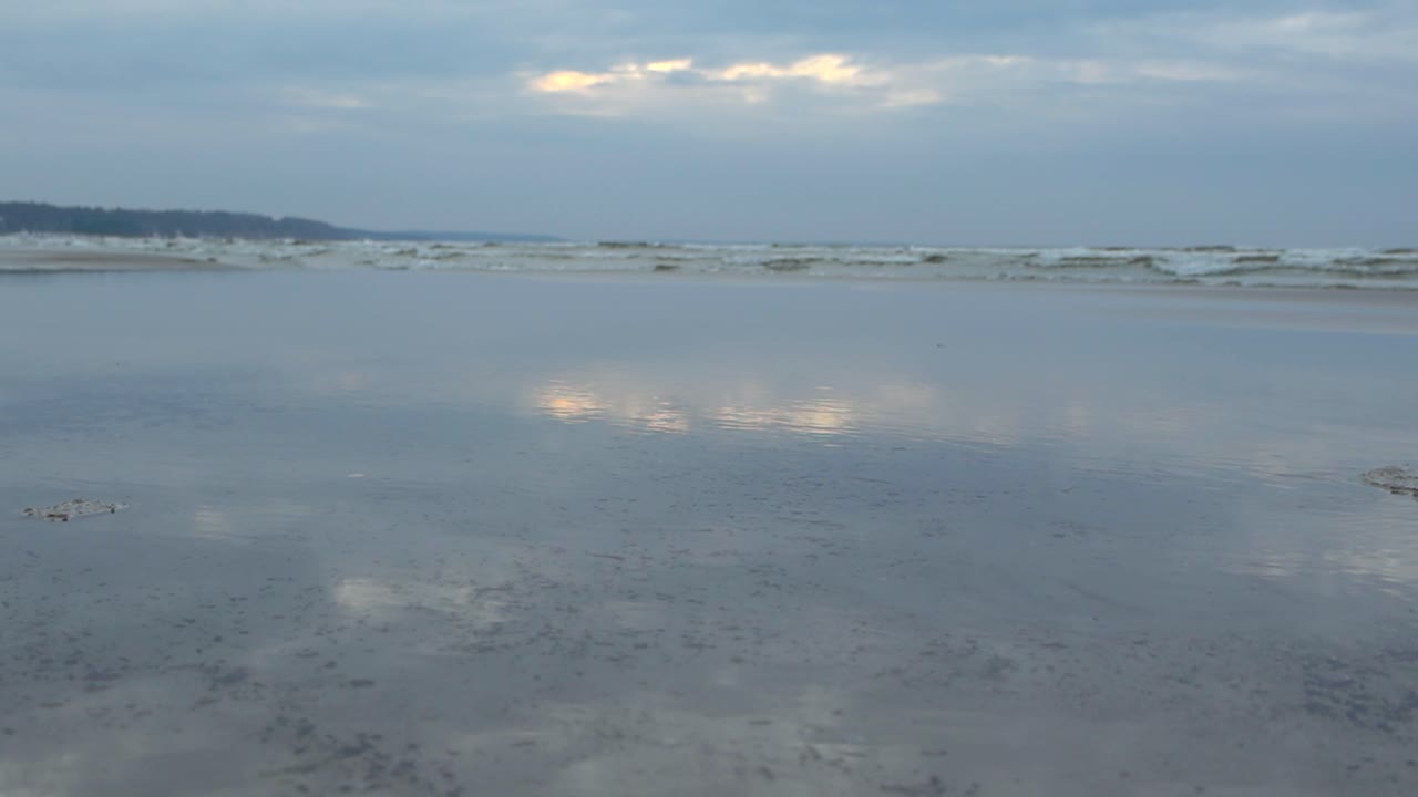 Gorgeous ocean sea water waves coming to a reflective sandy blue toned shoreline at a beach in Vääna during a cloudy day while the sun slightly glares and reflects on the still water in the foreground