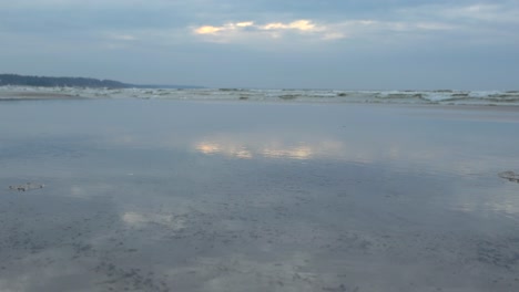 gorgeous ocean sea water waves coming to a reflective sandy blue toned shoreline at a beach in vääna during a cloudy day while the sun slightly glares and reflects on the still water in the foreground