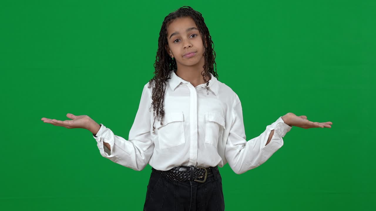 Unsure puzzled African American teen girl holding invisible on green screen shrugging shoulders looking at camera. Portrait of confused teenager posing at chromakey background. Choice concept.