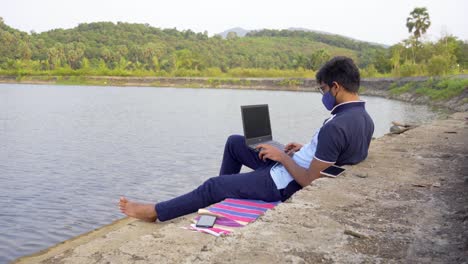 working remotely from laptop waring mask location lakeside india new normal wide shot relaxed