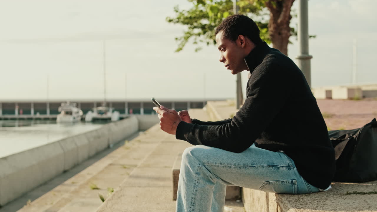 Young Man Sitting by Harbor and Using Mobile Phone