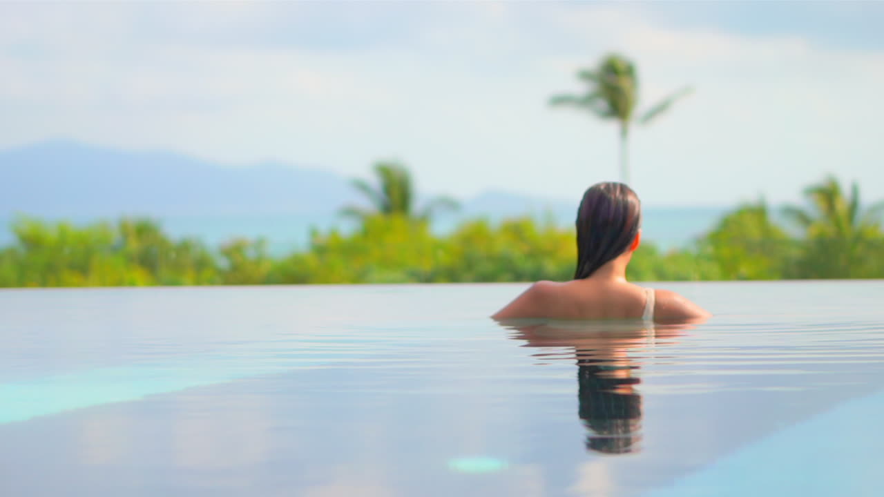 Rücken einer sinnlichen exotischen Frau in einem Infinity-Pool, die das Wasser genießt, mit Blick auf eine tropische Landschaft an einem heißen Sommertag, Zeitlupe, Vollbild.
