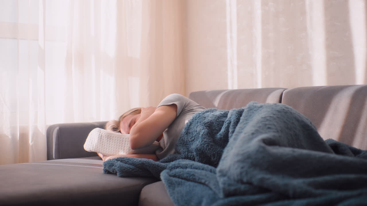 Exhausted banker collapsed on couch after long day, curling beneath soft blanket with phone beside her, hiding face behind pillow as early sunlight streams gently across peaceful room interior